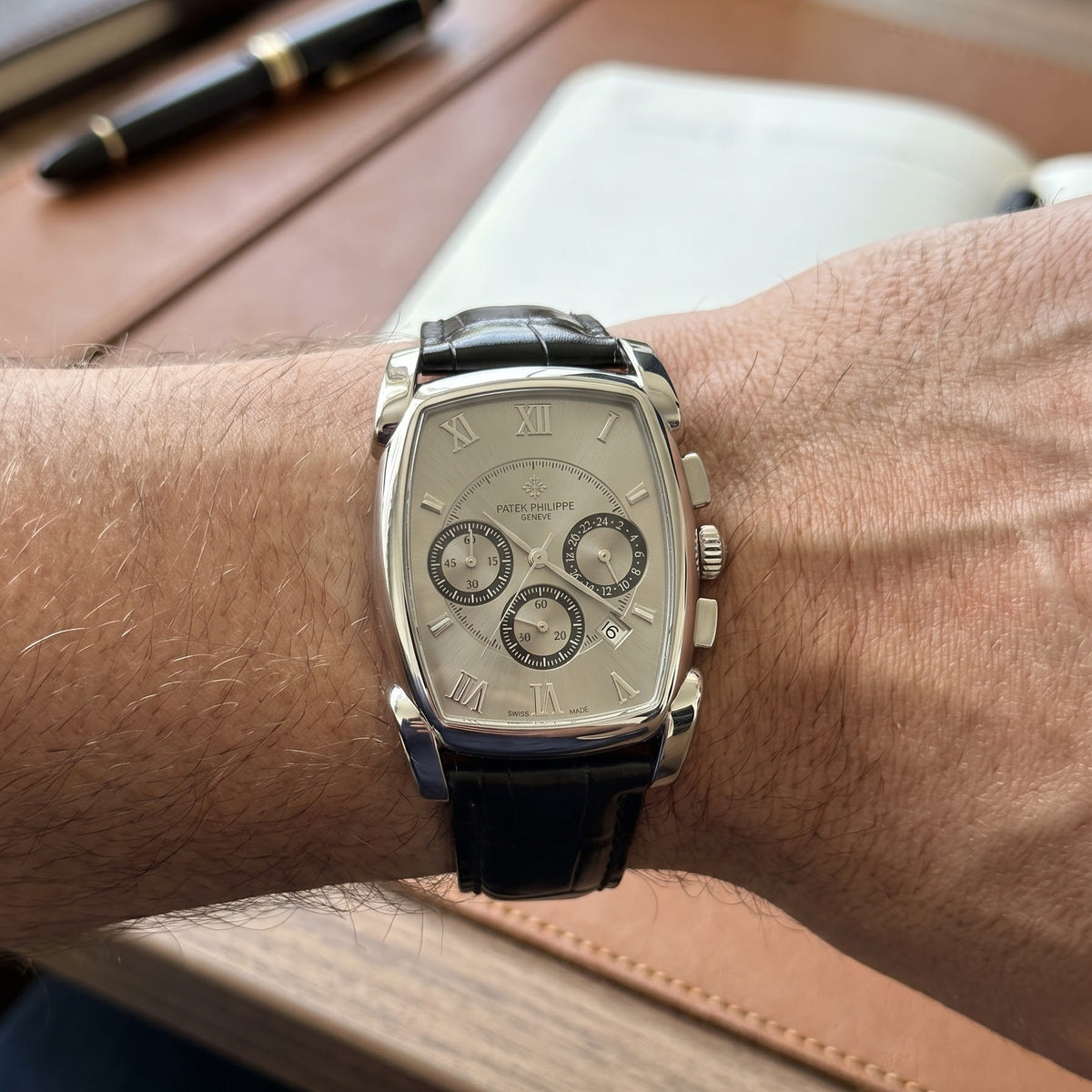 A rectangular silver-tone watch with a black leather strap and a multi-dial silver dial, worn on a person's wrist over a wooden desk background.