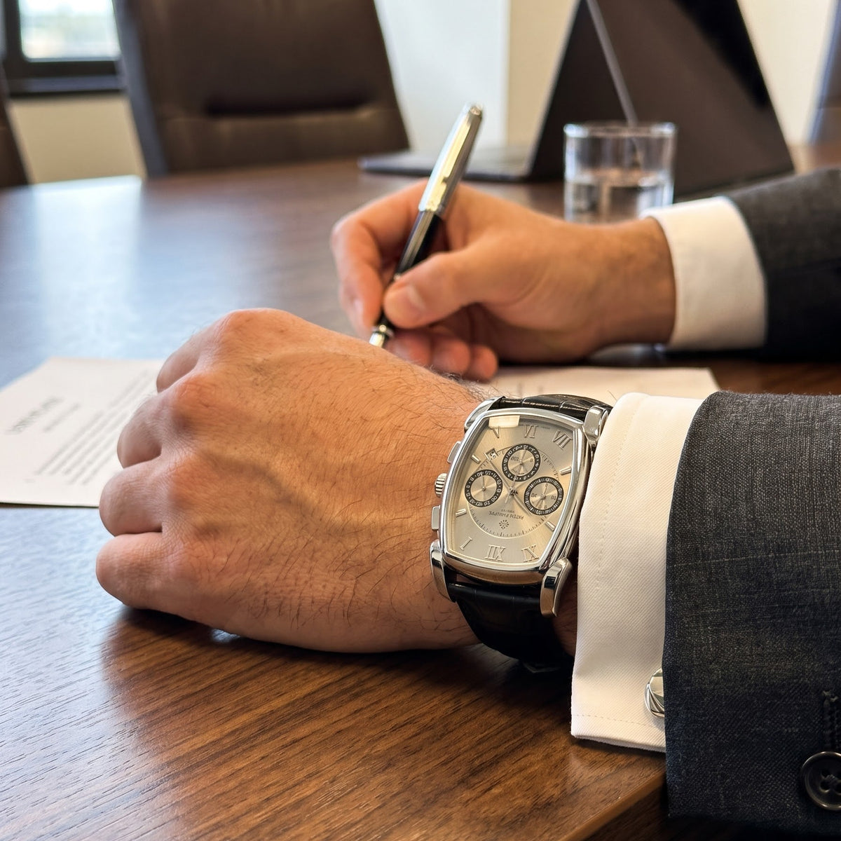 A luxury chronograph watch with a silver dial and black strap worn on a person's wrist, shown in a professional office setting with a wooden conference table, laptop, and pen.