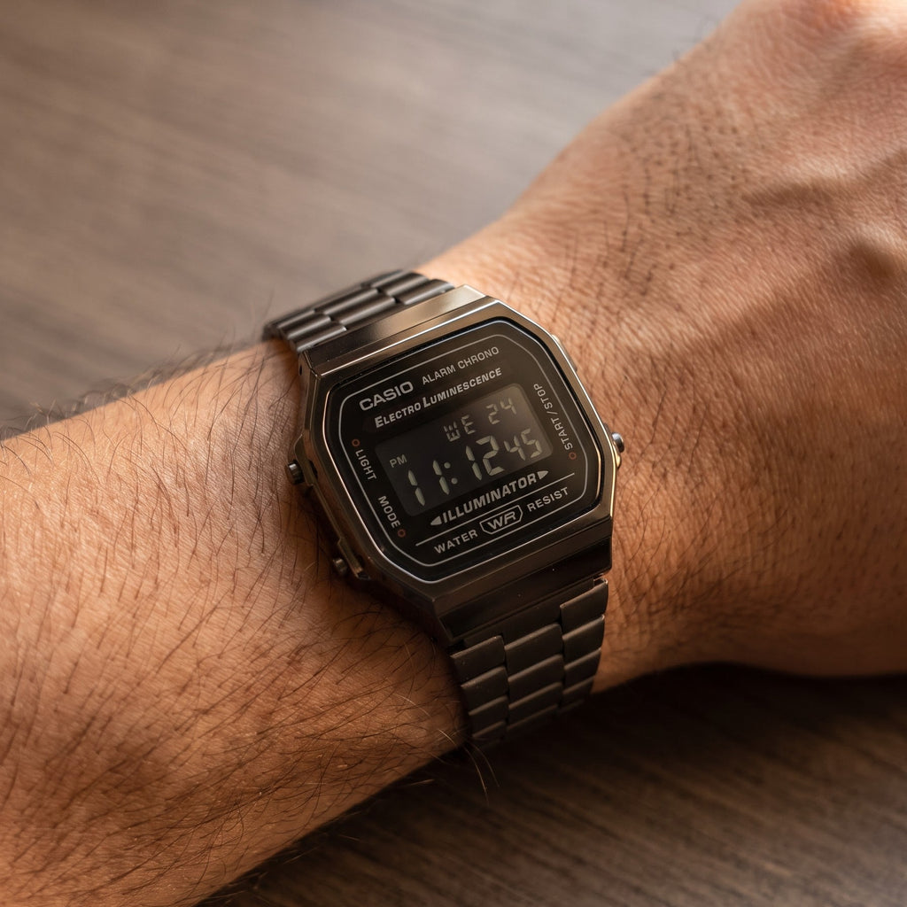 A Casio digital watch with a dark gunmetal metallic link band and matching case, worn on a person's wrist against a dark wood background.