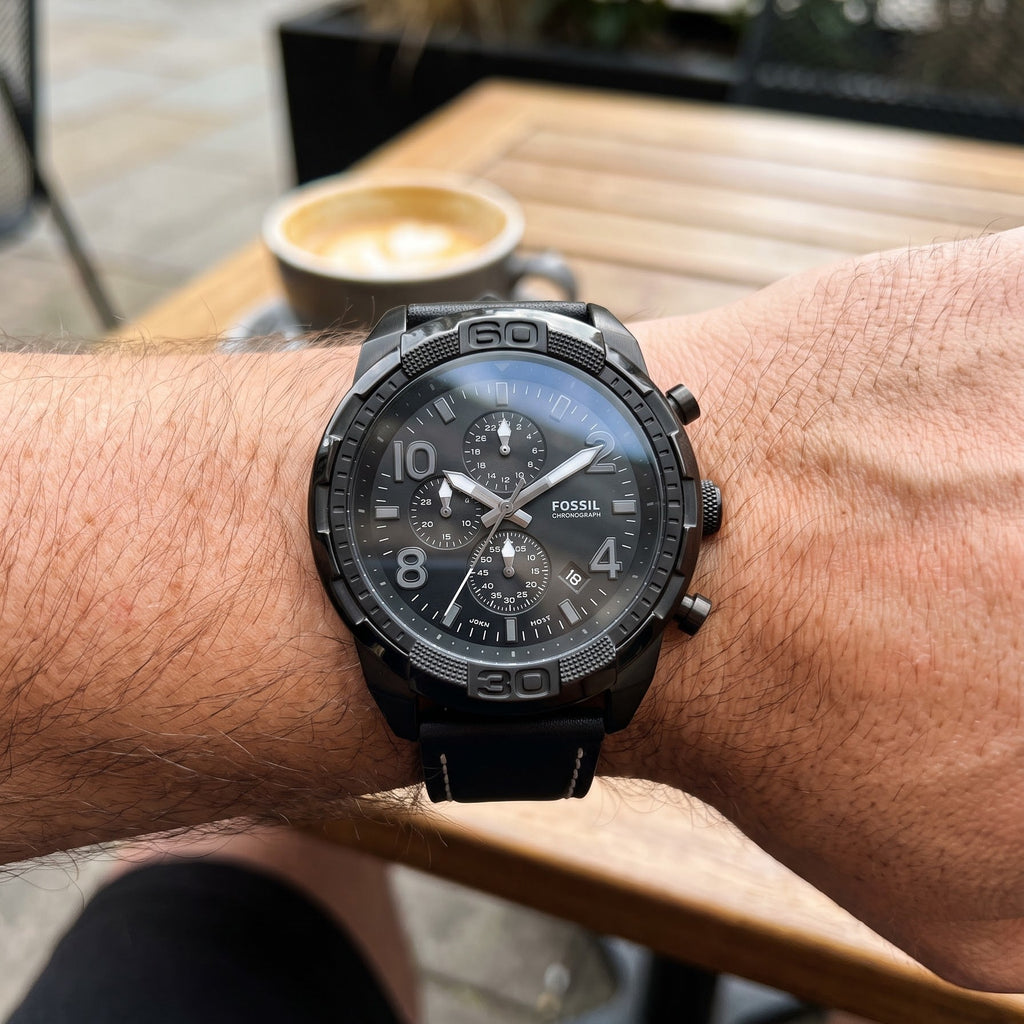 A close-up of a black Fossil chronograph watch with a black strap worn on a person's wrist, set against an outdoor cafe background with a wooden table and a cup of coffee.