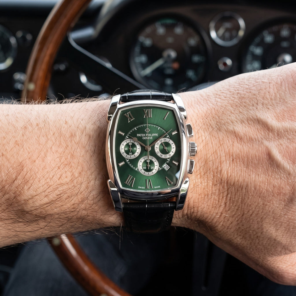 A wrist-worn chronograph watch with a green dial and silver-toned case, shown on a person's wrist in front of a wooden steering wheel and car dashboard.